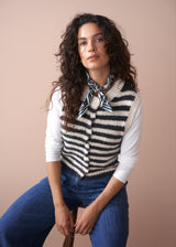 Woman wearing a striped vest over a white shirt with blue jeans against a beige background