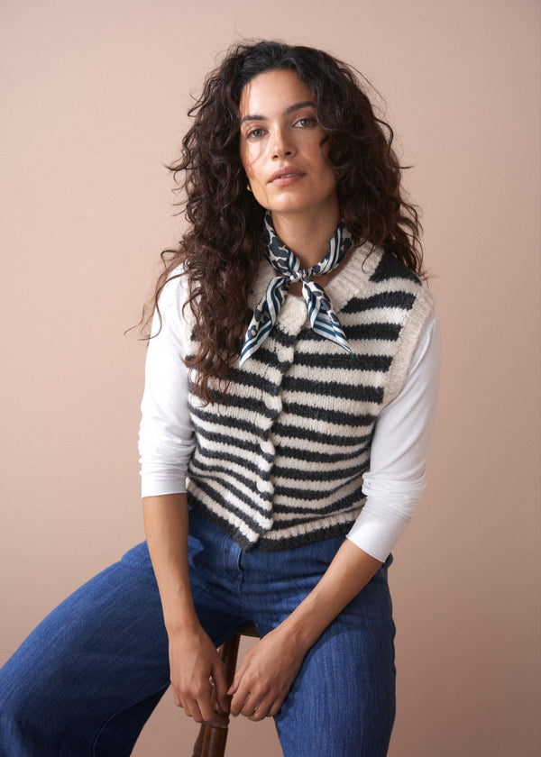 Woman wearing a striped vest over a white shirt with blue jeans against a beige background