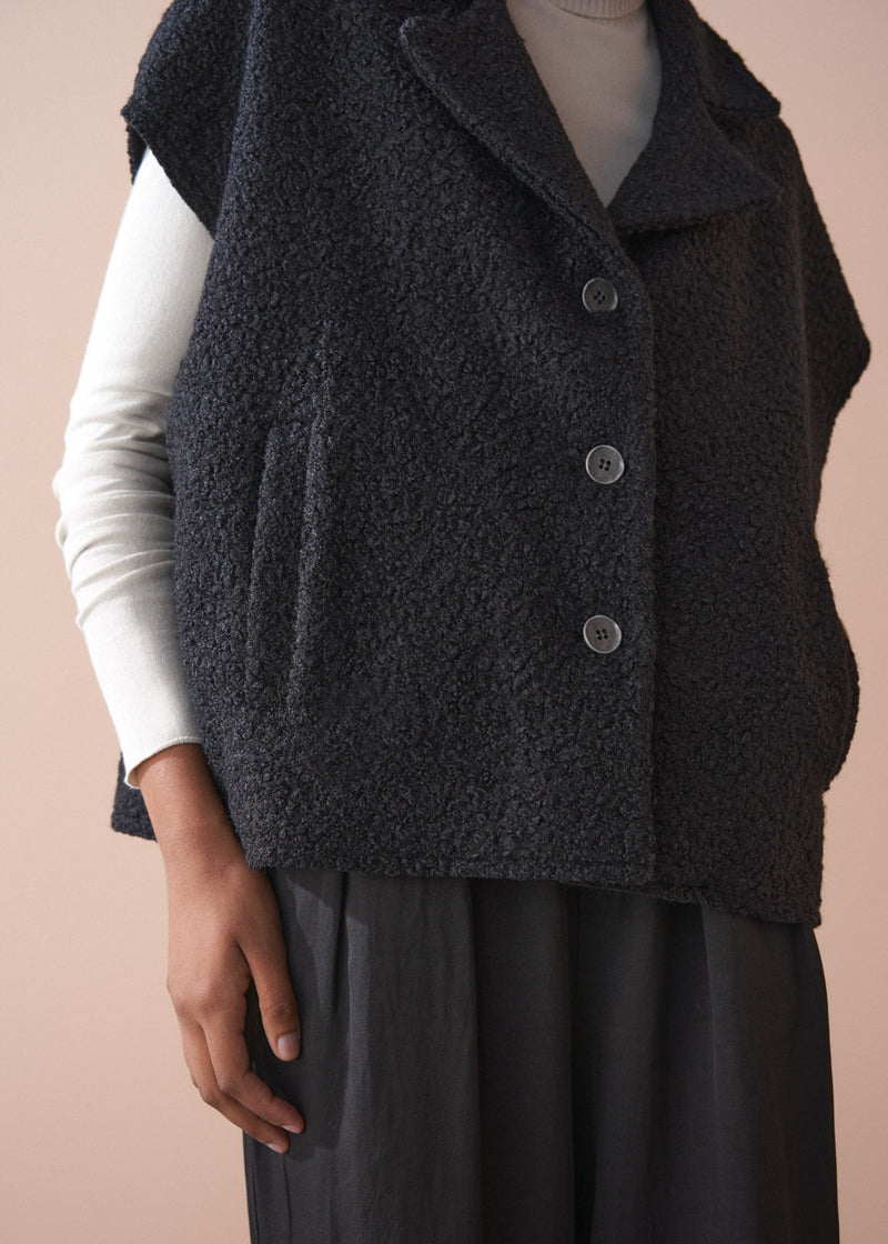 Person wearing a black boucle gilet jacket over a cream poloneck and black trousers against a beige background