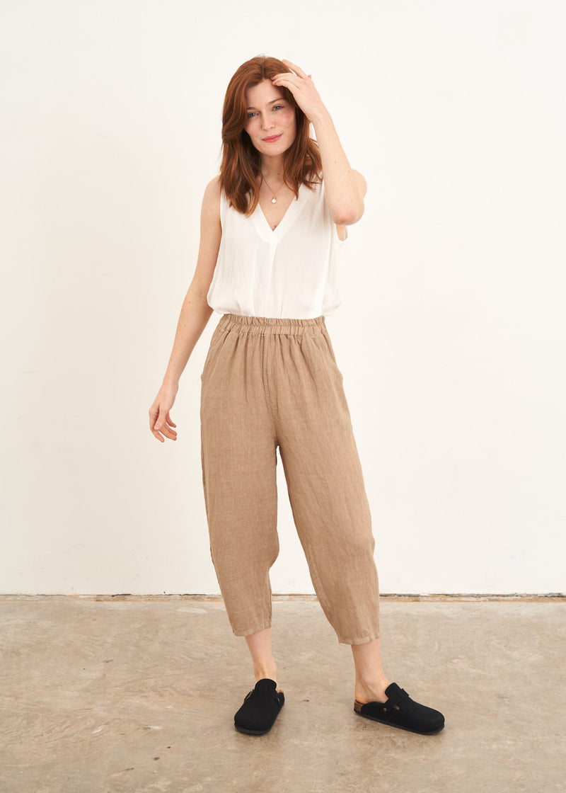 A model wearing a pair of camel cropped linen trousers with a white sleeveless top and black clogs