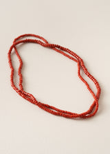 A long strand necklace made of small red cylindrical beads, arranged in three overlapping loops on a light beige textured background.
