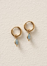 A pair of gold huggie hoop earrings with dangling light blue teardrop gemstones, displayed on a textured off-white background.