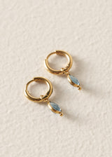 A pair of gold huggie hoop earrings with dangling marquise-cut light blue gemstones, displayed on a textured off-white surface.