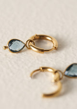 Gold huggie hoop earrings with a teardrop-shaped light blue gemstone, shown on a textured off-white background.