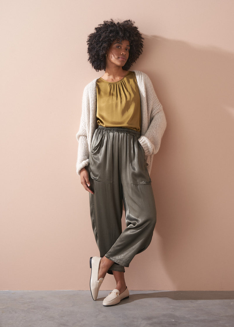 Woman wearing a gold satin top and green satin trousers against a beige wall