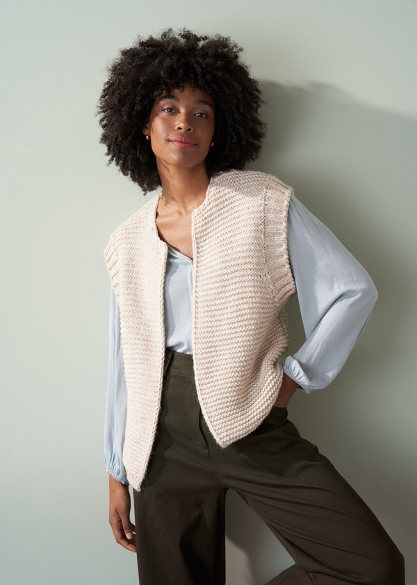 Woman wearing a beige knitted vest over a pale blue shirt and dark green trousers with a neutral background