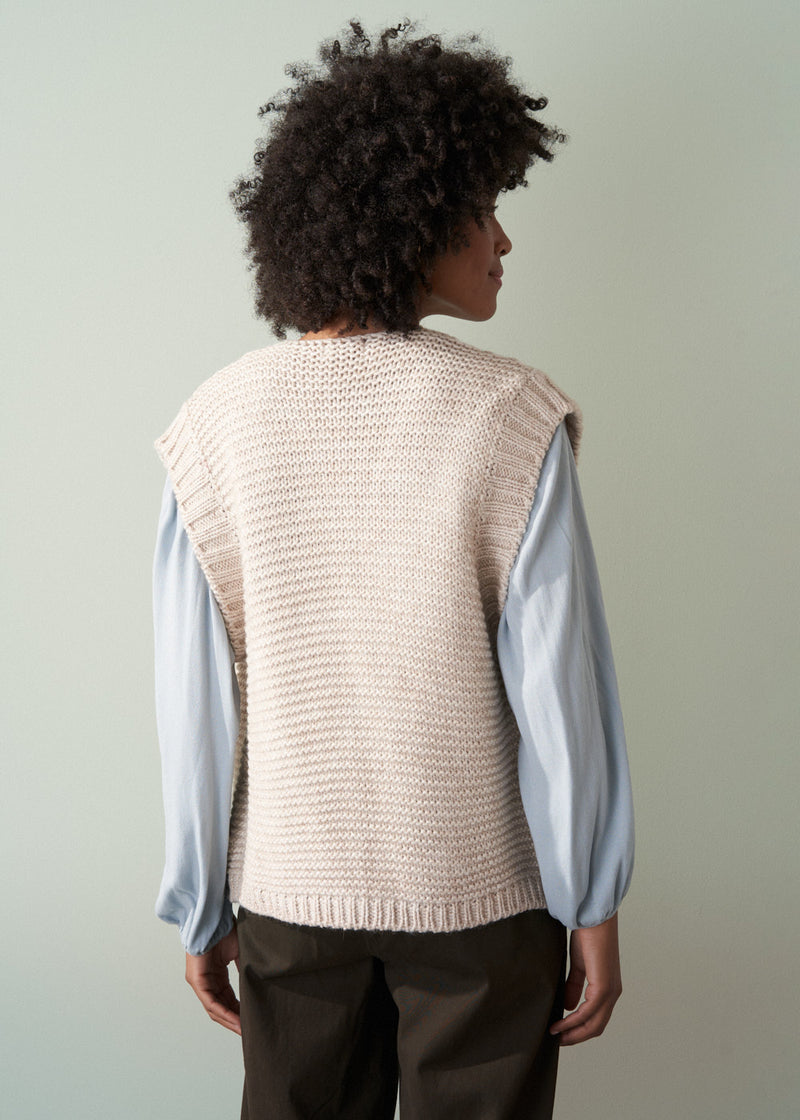 Woman wearing a beige knitted vest over a pale blue shirt and dark green trousers with a neutral background