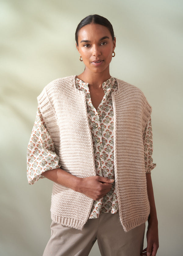 Woman wearing a beige knitted cardigan over a floral cotton blouse against a plain background