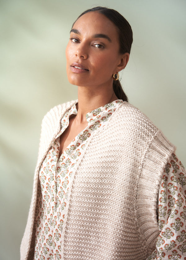 Woman wearing a beige knitted cardigan over a floral cotton blouse against a plain background