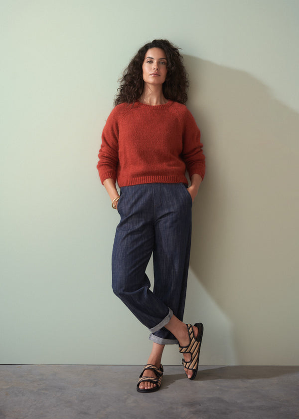 Woman wearing a rust-colored sweater and dark blue jeans against a plain wall.