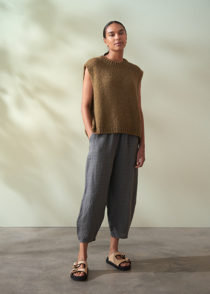 Woman wearing a khaki green sleeveless sweater and checkered pants against a neutral background