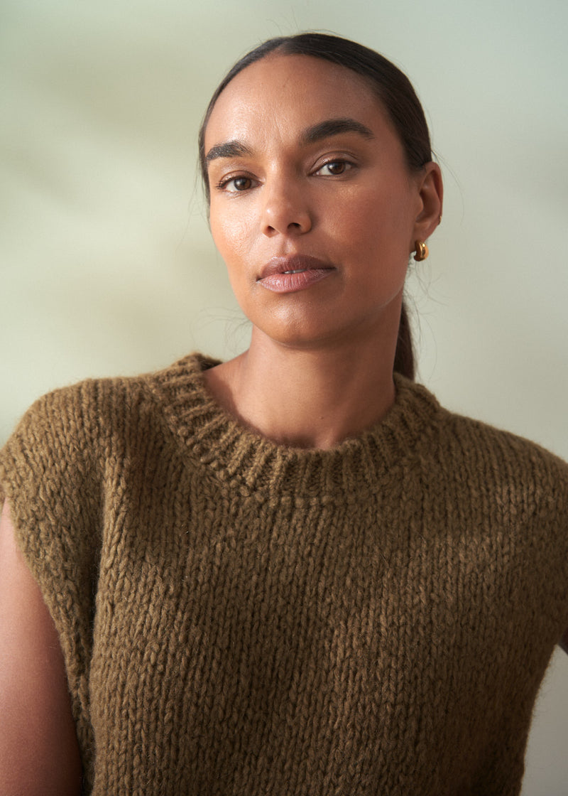 Woman wearing a khaki green knitted sleeveless sweater against a plain background