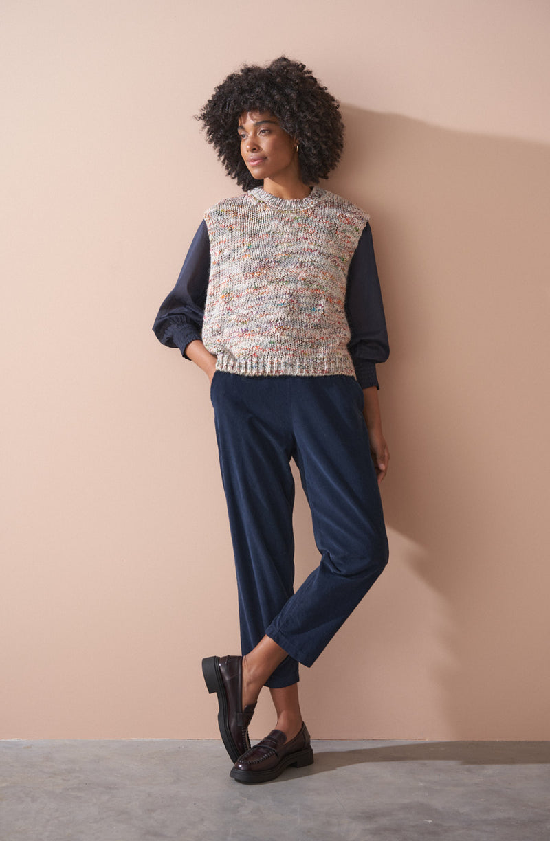 Woman wearing a sleeveless space dye sleeveless top and blue pants against a beige wall.