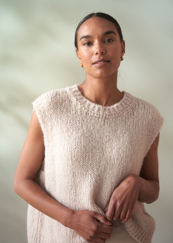 Woman wearing a beige knitted sleeveless sweater against a plain background