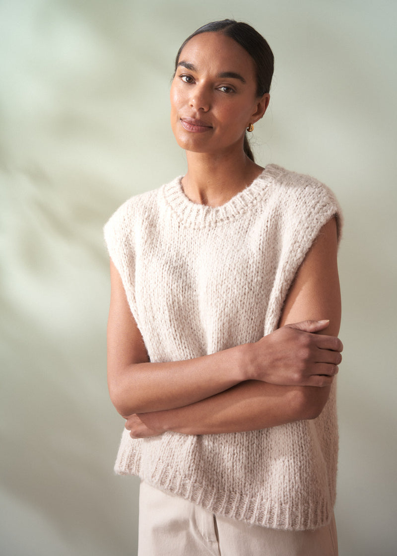 Woman wearing a beige knitted sleeveless top against a plain background