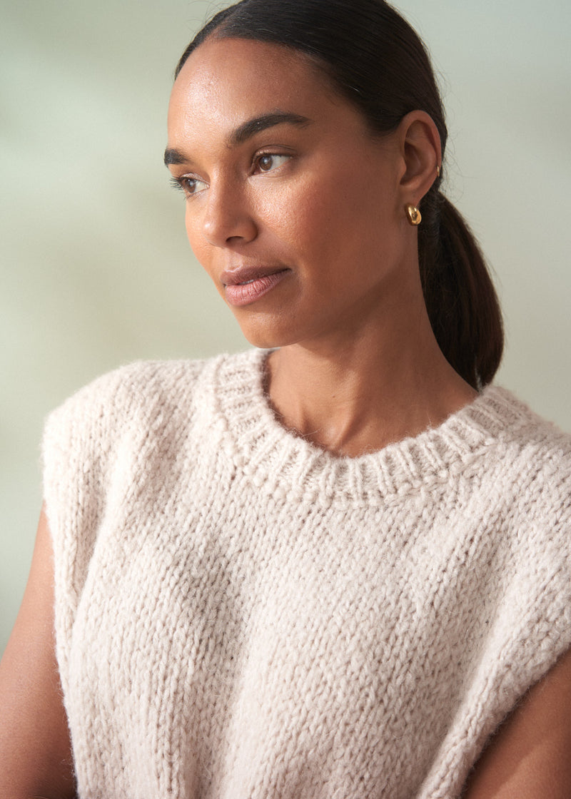 Woman wearing a beige sleeveless knitted top against a plain background