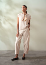 Woman wearing a light-colored sleeveless top and trousers with a scarf, standing against a neutral background.