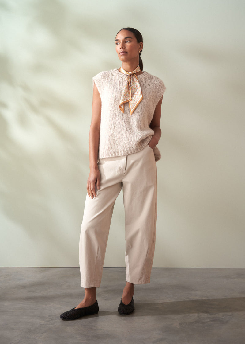 Woman wearing a light-colored sleeveless top and trousers with a scarf, standing against a neutral background.