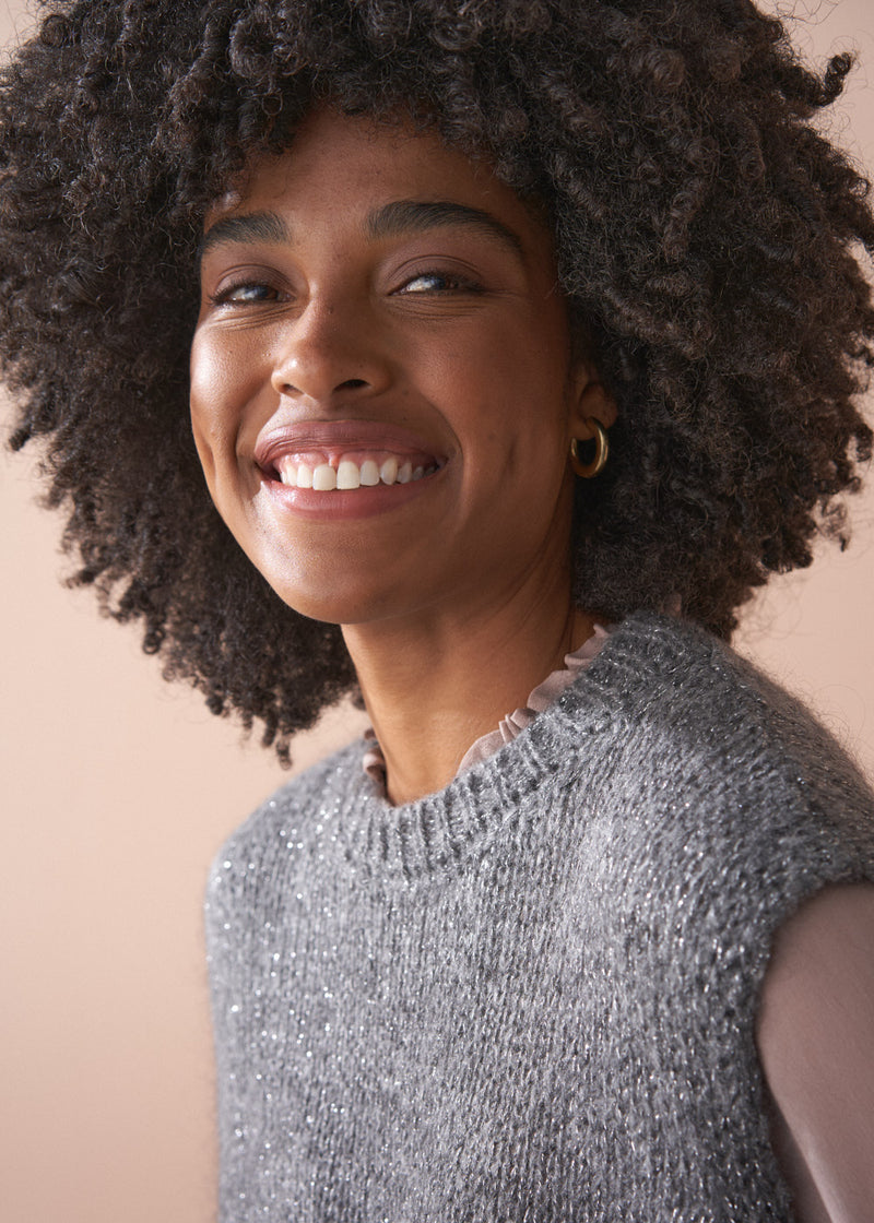Woman wearing a grey sleeveless sparkle sweater against a beige background