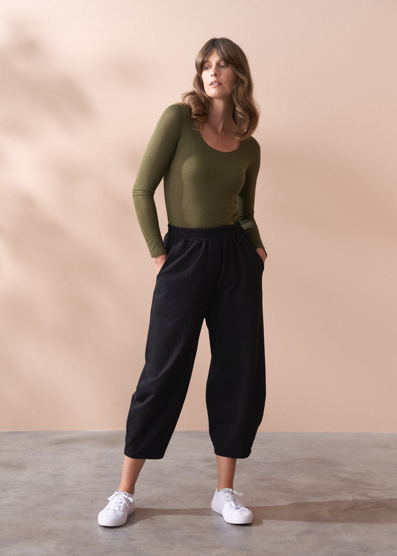 Woman wearing a green top and black barrel leg cotton trousers against a beige background