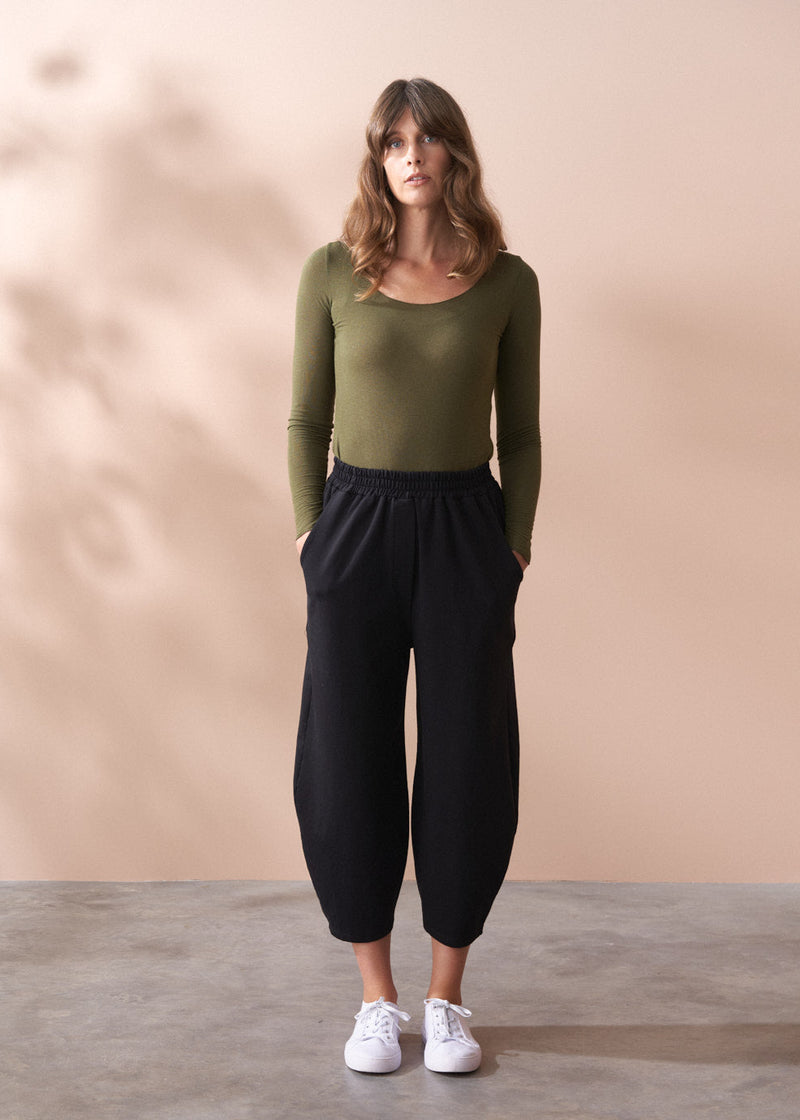 Woman wearing a green top and black barrel leg cotton trousers against a beige background