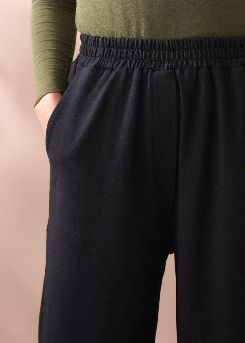 Person wearing black barrel leg cotton trousers and a green top against a beige background