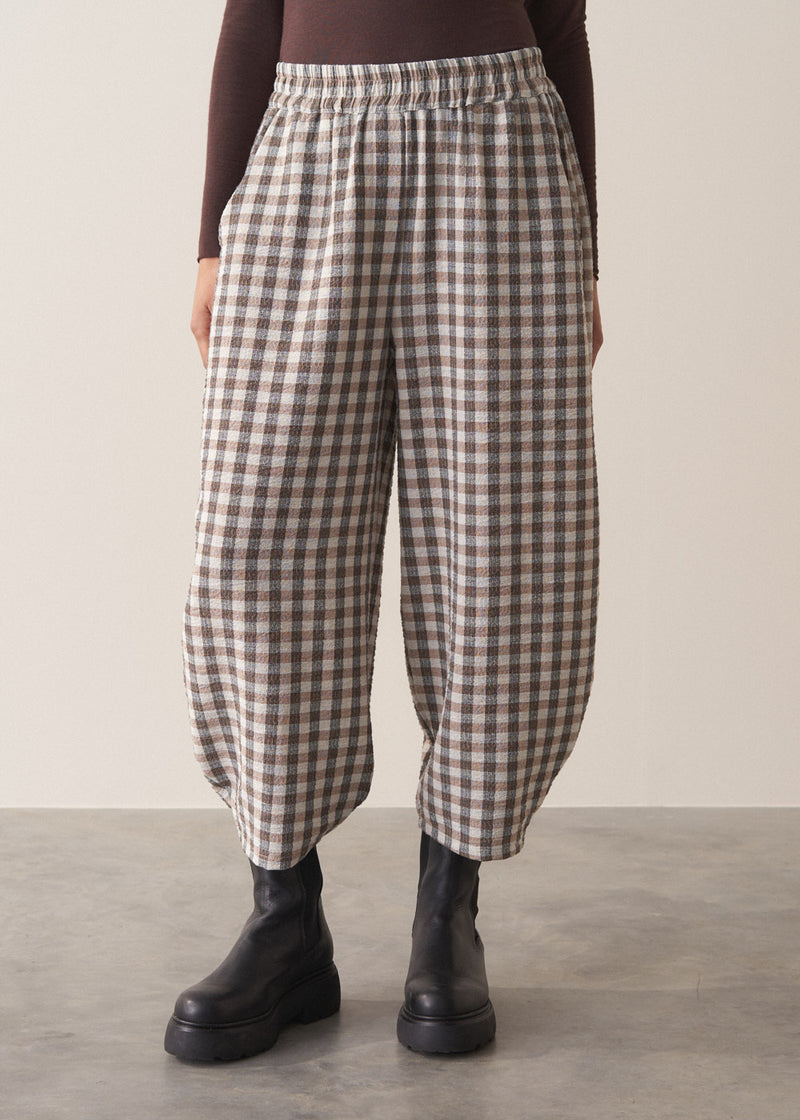 Woman wearing a brown poloneck sweater and checked barrel leg trousers against a plain background