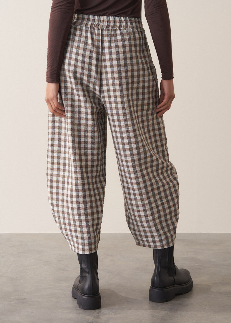 Woman wearing a brown poloneck sweater and checked barrel leg trousers against a plain background