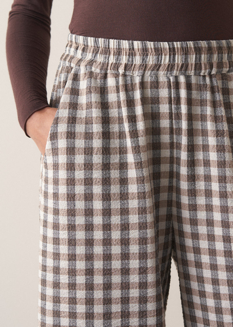 Woman wearing a brown poloneck sweater and checked barrel leg trousers against a plain background