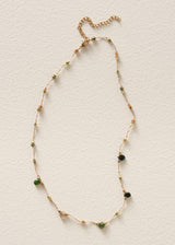 A delicate gold chain necklace with small green and orange beads, accented by larger green and black beads, laid flat on a textured off-white background.