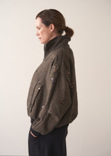 Person wearing a brown textured bomber jacket with sequin detail and black trousers on a plain background