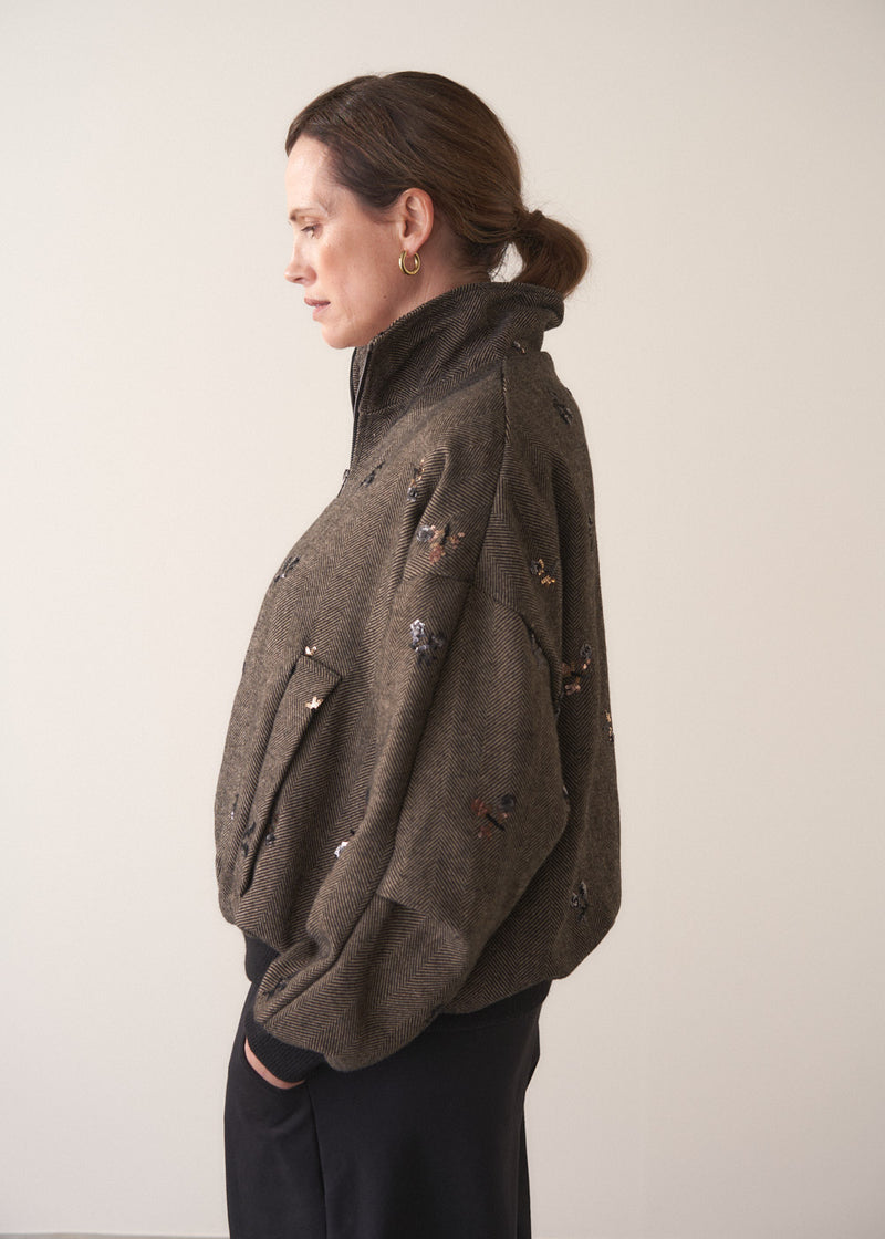 Person wearing a brown textured bomber jacket with sequin detail and black trousers on a plain background
