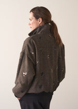 Person wearing a brown textured bomber jacket with sequin detail and black trousers on a plain background