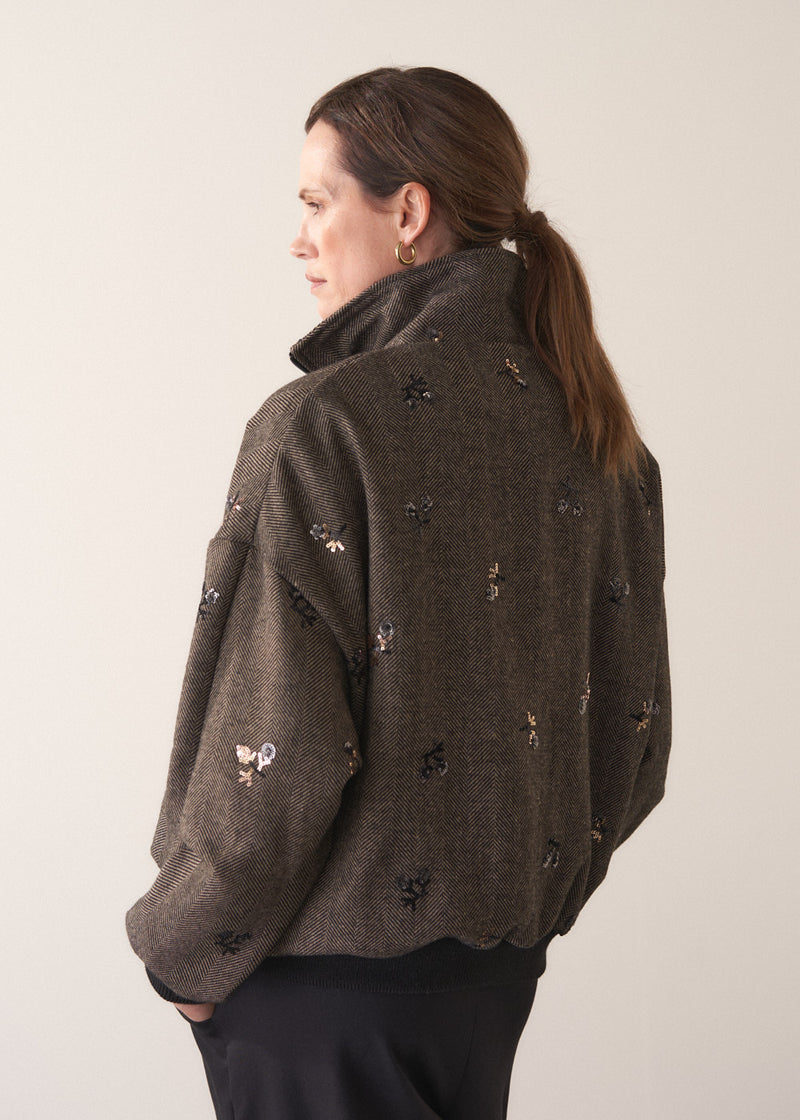 Person wearing a brown textured bomber jacket with sequin detail and black trousers on a plain background