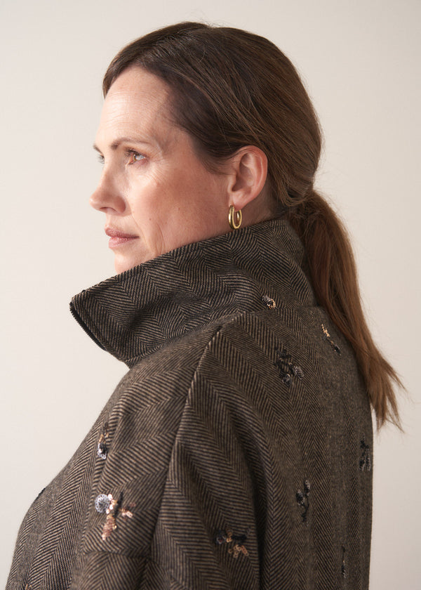 Person wearing a brown textured bomber jacket with sequin detail and black trousers on a plain background