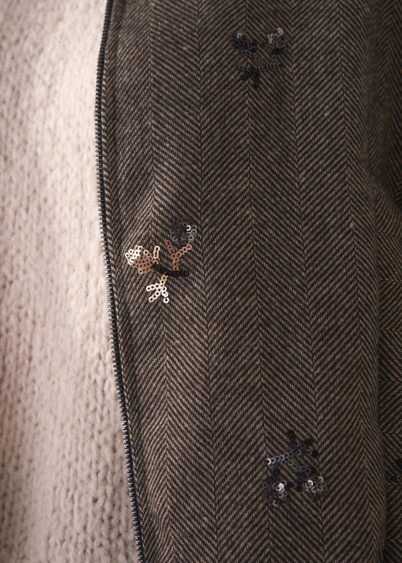 Person wearing a brown textured bomber jacket with sequin detail and black trousers on a plain background