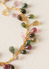 A close-up of a delicate gold chain necklace draped diagonally, featuring small teardrop-shaped faceted gemstones in pink, green, yellow, and dark brown, wrapped individually in gold wire on a textured off-white background.