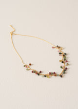 A delicate gold chain necklace featuring small, irregular multicolored gemstone chips in shades of green, pink, and red, displayed on a textured off-white background.