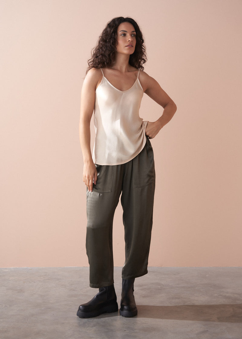Woman wearing a beige satin camisole top and dark green trousers against a plain background
