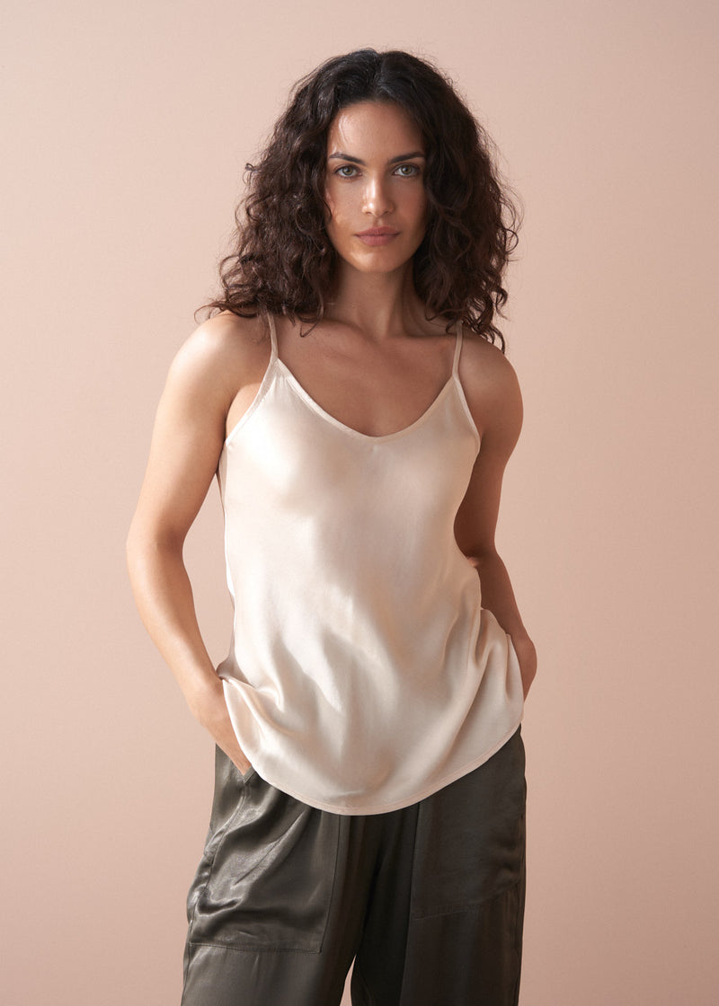 Woman wearing a beige satin camisole top and dark green trousers against a plain background