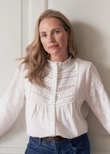 A woman with blonde hair wearing a white long-sleeve blouse with intricate lace and embroidery on the chest and sleeves, paired with blue jeans, leaning against a white paneled wall.