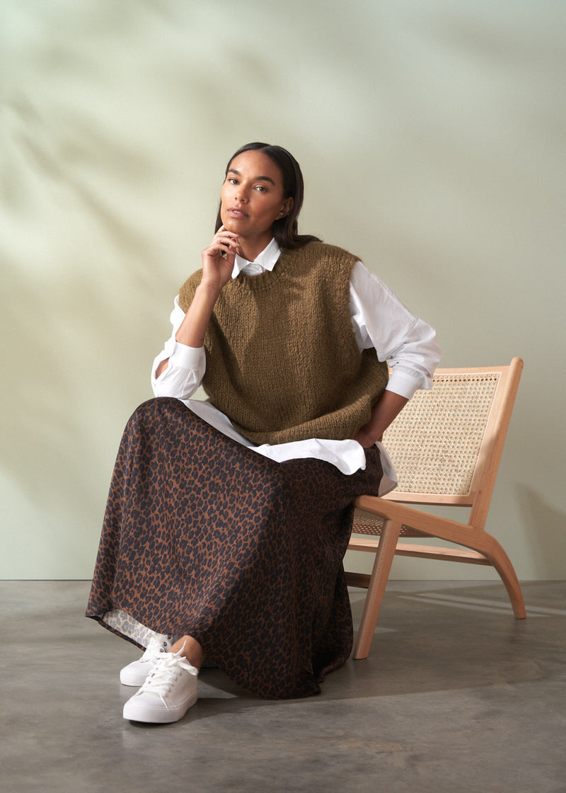 Woman sitting on a chair wearing a brown knitted vest over a white shirt with a leopard print skirt.