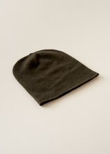 A dark olive green knit beanie hat, photographed from an angle on a clean, light beige background.