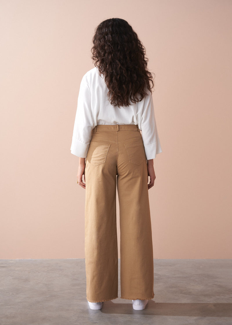 Woman wearing a white blouse and tan wide-leg pants against a beige wall.