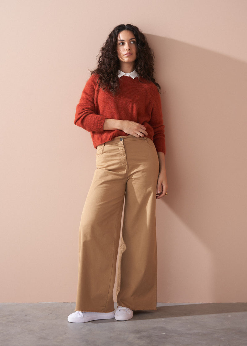 Woman wearing a red sweater and beige pants against a beige wall