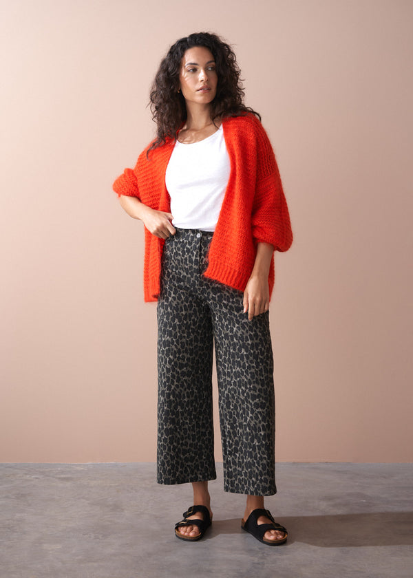 Woman wearing a red cardigan, white top, and leopard print trousers against a beige background