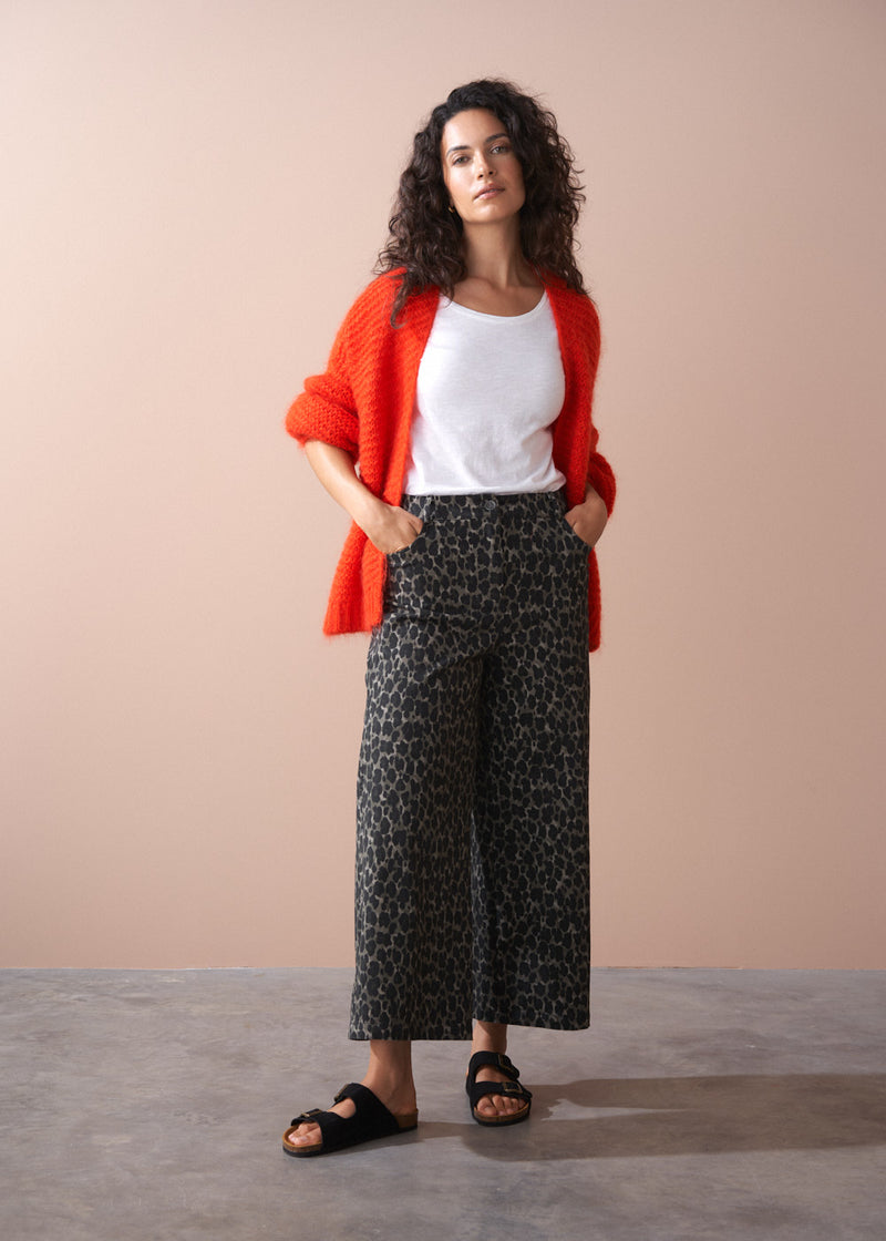 Woman wearing a red cardigan, white top, and leopard print trousers against a beige background