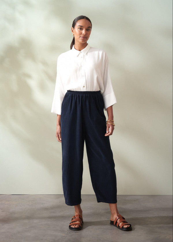 Woman wearing a white shirt and navy corduroy barrel leg trousers against a plain background
