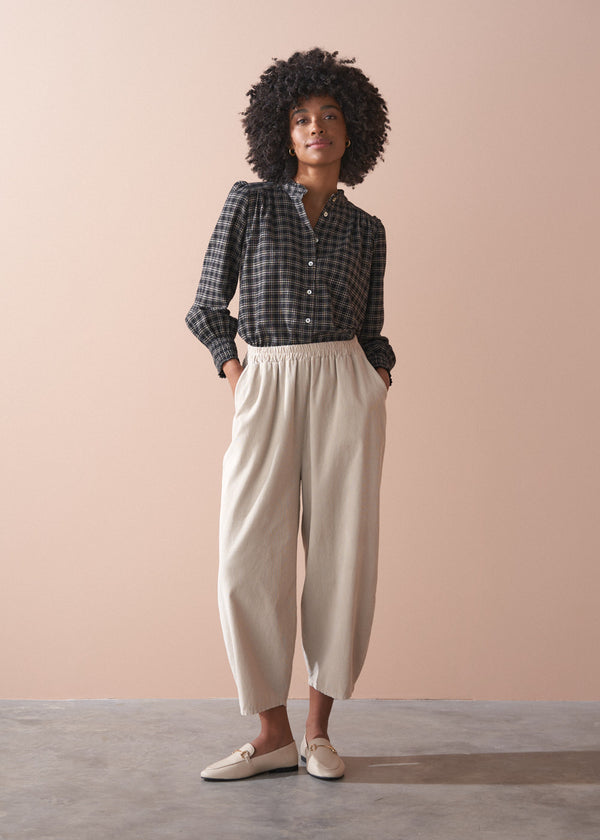 Woman wearing a check shirt and beige corduroy barrel leg trousers against a beige background