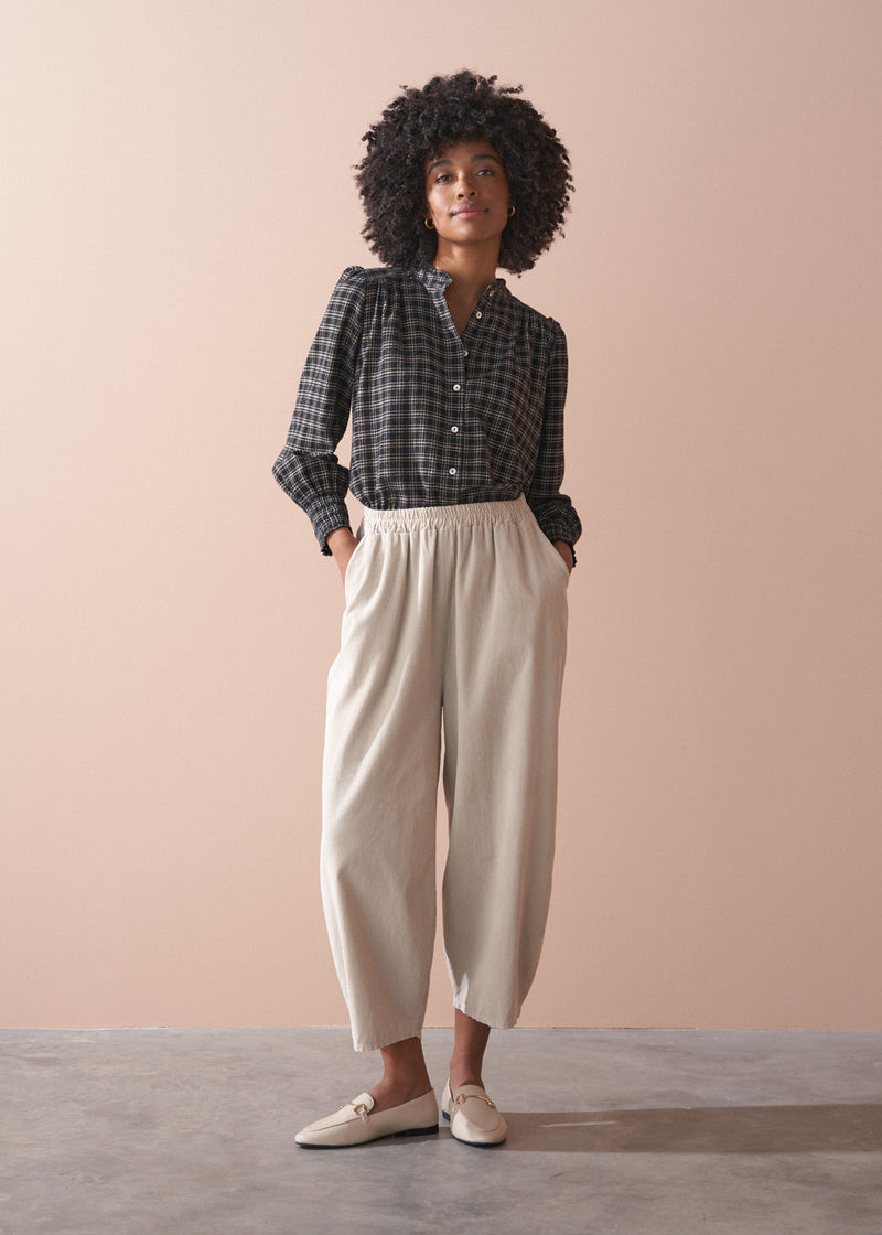Woman wearing a check shirt and beige corduroy barrel leg trousers against a beige background